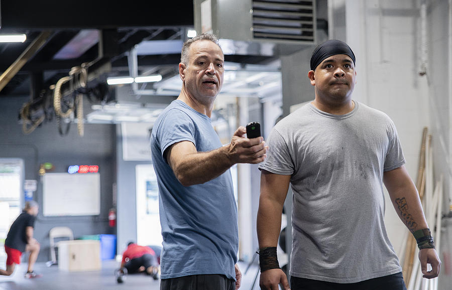 The senior Latino man, the coach, showing the fitness results to the young Hispanic athlete, powerlifter, on a smart fitness device in the gym during the workout. Photograph by Alex Potemkin