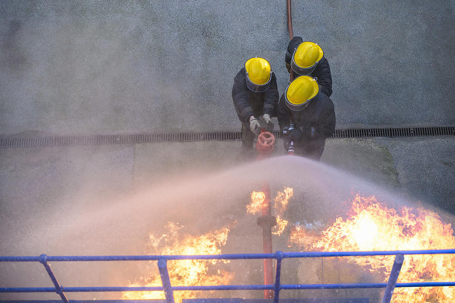 Three firefighters putting out fire in fire simulation training facility, overhead view Photograph by Monty Rakusen