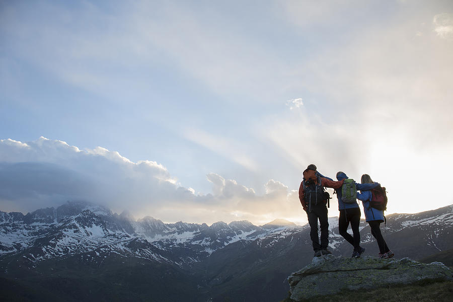 Three hikers link arms on rock above mountains Photograph by Ascent Xmedia
