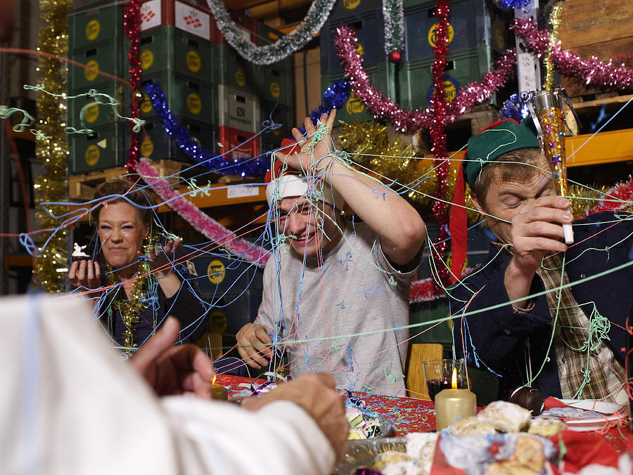 Three people covered in party streamers laughing at charismas table in warehouse Photograph by Ulrik Tofte