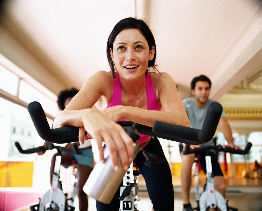Three people sitting on exercising bikes in gym, close-up Photograph by Commercial Eye