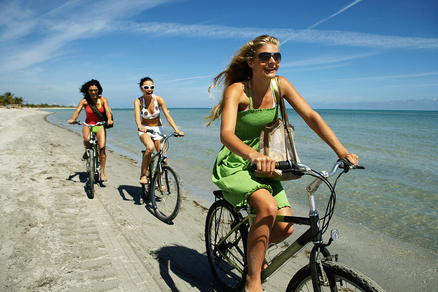 Three young women riding bicycles on beach, smiling Photograph by Kraig Scarbinsky