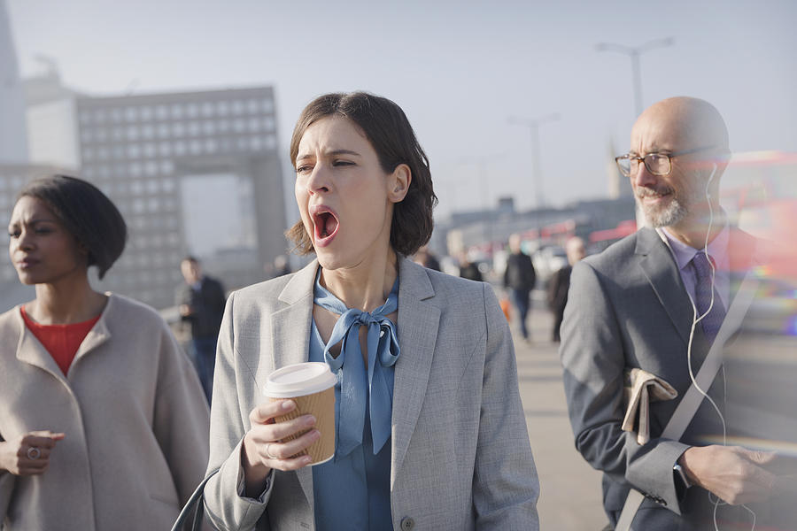 Tired businesswoman with coffee yawning on sunny morning urban pedestrian bridge Photograph by Caia Image