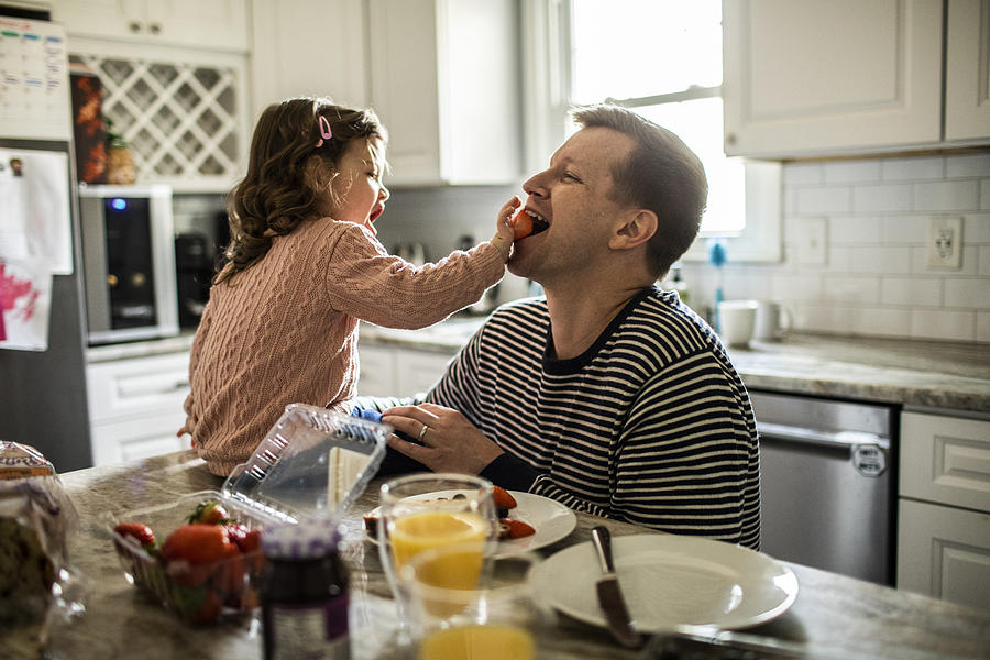 Toddler girl feeding her father a strawberry in kitchen Photograph by MoMo Productions