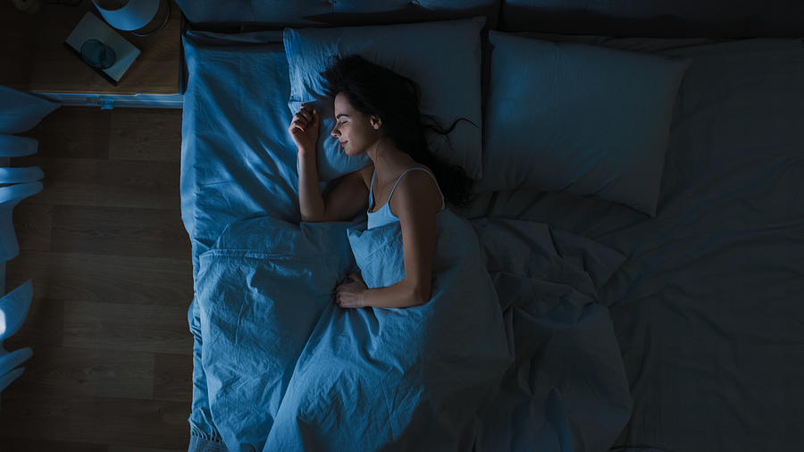 Top View of Beautiful Young Woman Sleeping Cozily on a Bed in His Bedroom at Night. Blue Nightly Colors with Cold Weak Lamppost Light Shining Through the Window. Photograph by Gorodenkoff