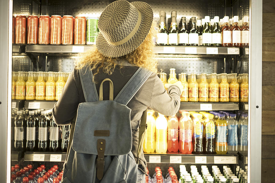 Travel woman viewed from back choosinf beverage in a fresh fridge - airport or station bar concept and traveler passenger choosing drinks - modern lifestyle backpack people buying drinks bottles Photograph by Simonapilolla