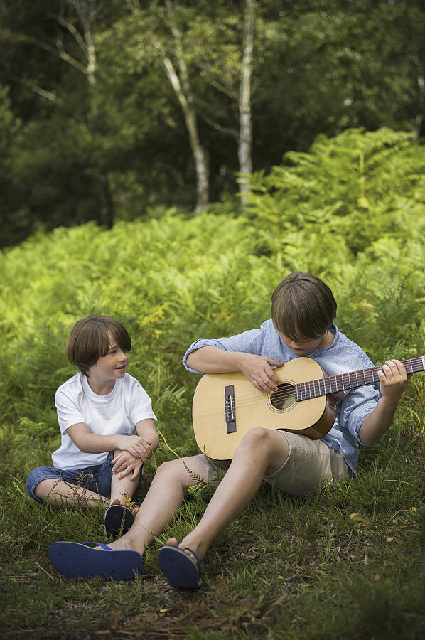 Two boys camping in New Forest. Sitting on the grass, one playing a guitar. Photograph by Mint Images - Emily Hancock