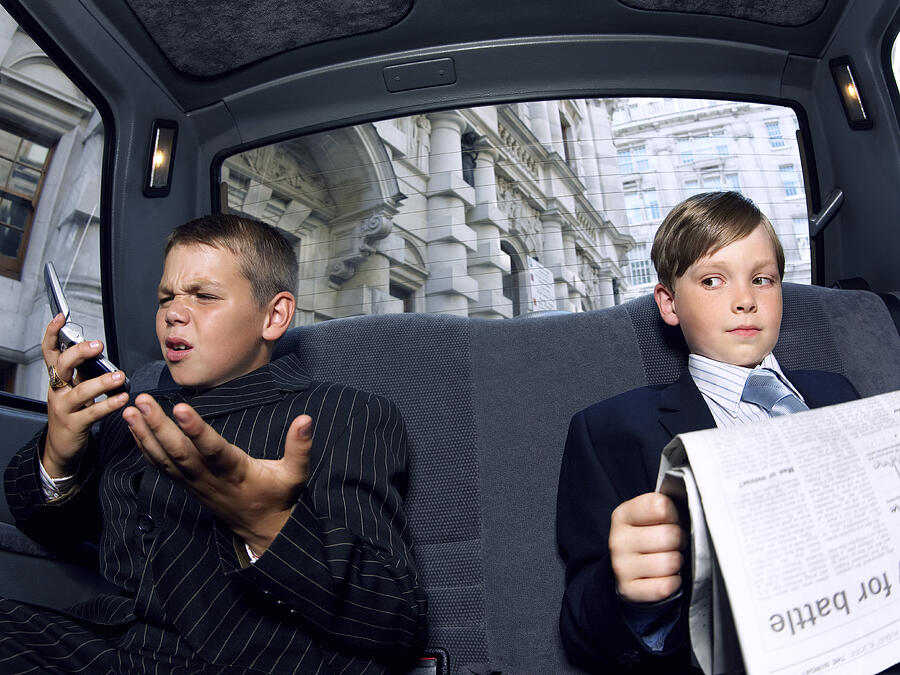 Two Boys Dressed as Businessmen in the back of a Taxi, one Reading a Newspaper the Other Looking Angry Using a Mobile Phone Photograph by Digital Vision.