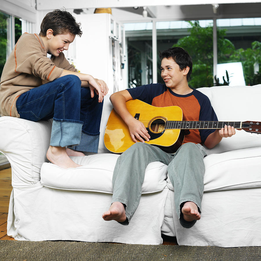 Two Brothers Laughing And Playing Guitar Sitting On A Sofa Photograph by Stockbyte