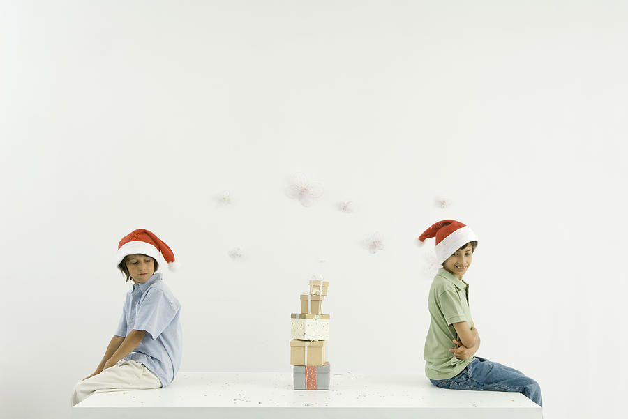 Two brothers sitting back to back, Christmas gifts stacked between them, both wearing Santa hats Photograph by PhotoAlto/Odilon Dimier