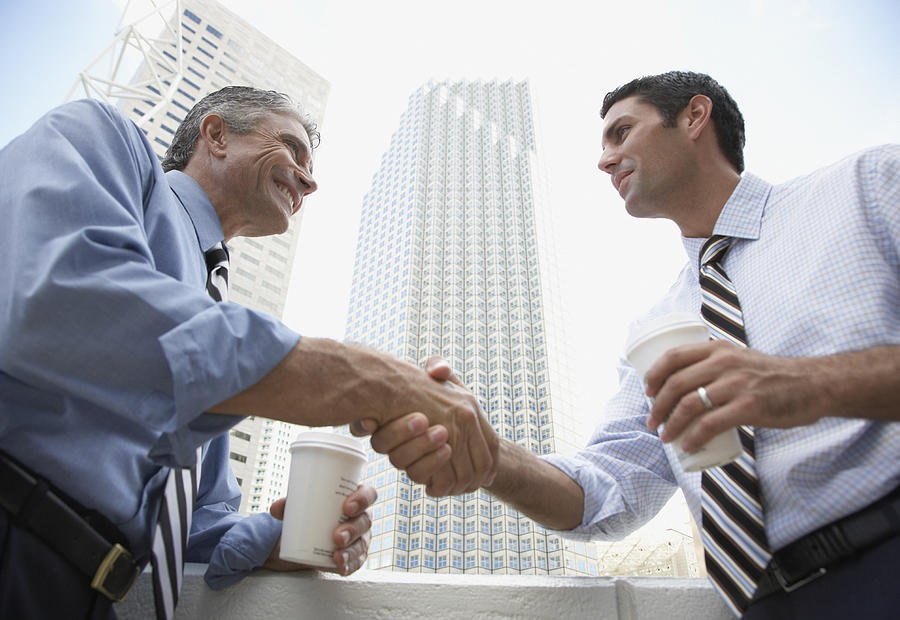 Two businessmen outdoors on a balcony shaking hands holding coffee cups Photograph by Tom Merton