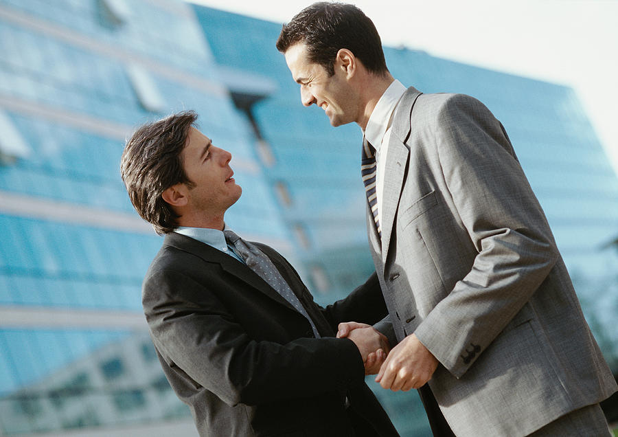Two businessmen shaking hands, buildings in background Photograph by Eric Audras