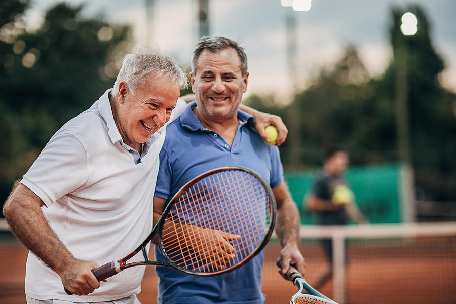 Two cheerful senior men talking while walking on the outdoor tennis court Photograph by South_agency