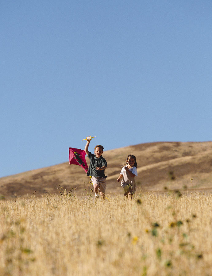 Two children run through a golden field with a kite Photograph by Photodisc