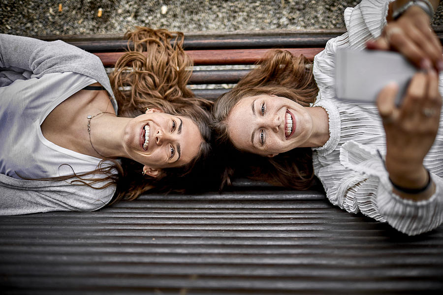 Two happy young women lying on a bench using cell phone Photograph by Oliver Rossi