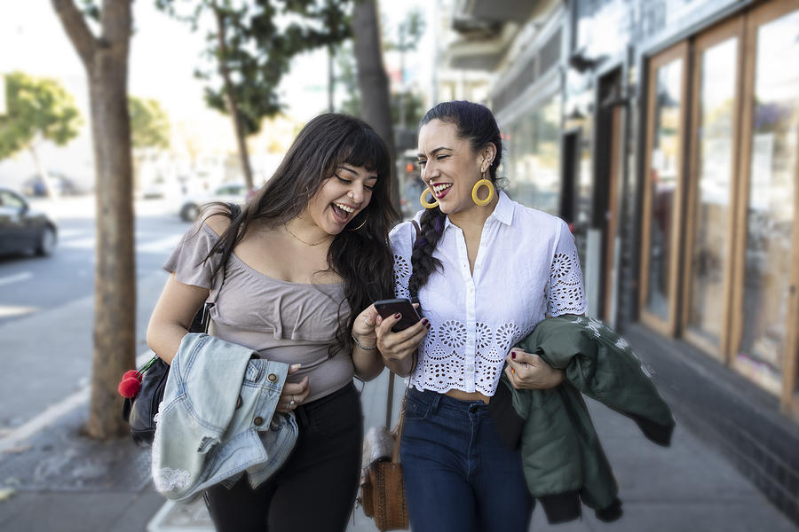 Two Latina Girlfriends Walking Along Sidewalk, Looking at Smart Phone, Laughing Photograph by Justin Lewis