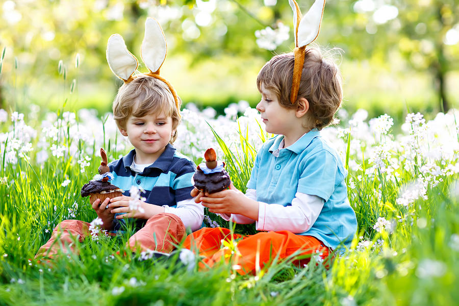 Two little boy friends in Easter bunny ears eating chocolate cakes and muffins Photograph by Romrodinka