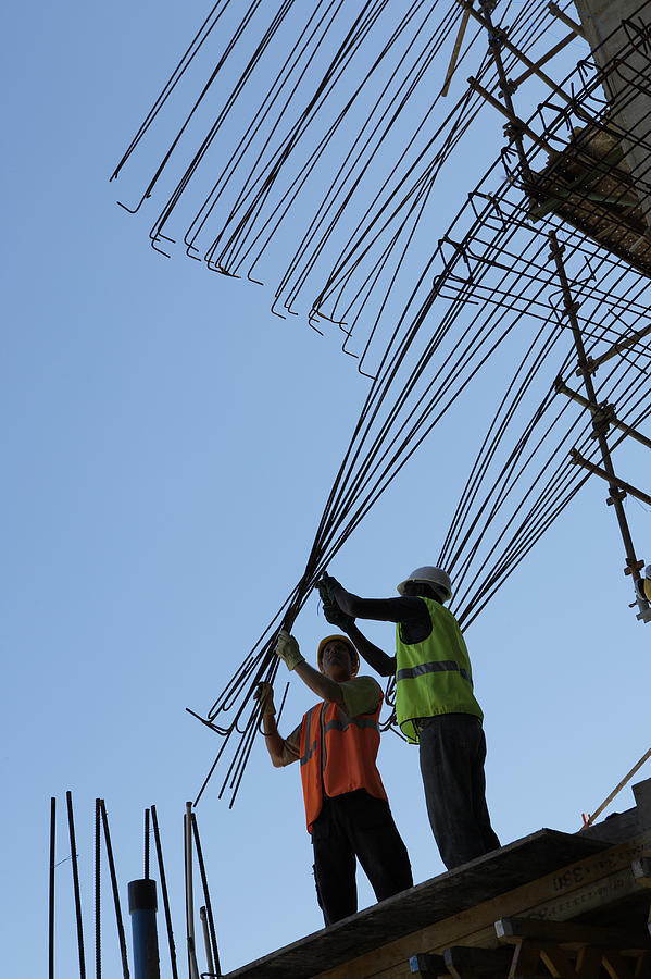 Two Male Construction Workers Stand on a Roof, Bending a Frame Into Position Photograph by Digital Vision.