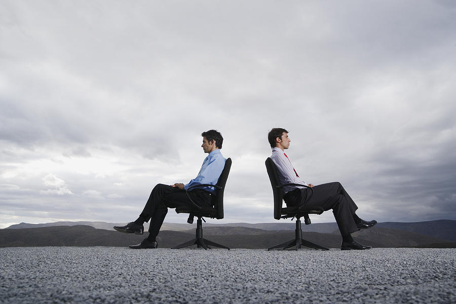 Two men sitting in office chairs outdoors with their backs against one another Photograph by OJO Images