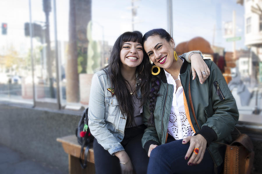 Two Millennial Latina Women Lean Into Each Other, Smiling, While Sitting on a Bench Photograph by Justin Lewis