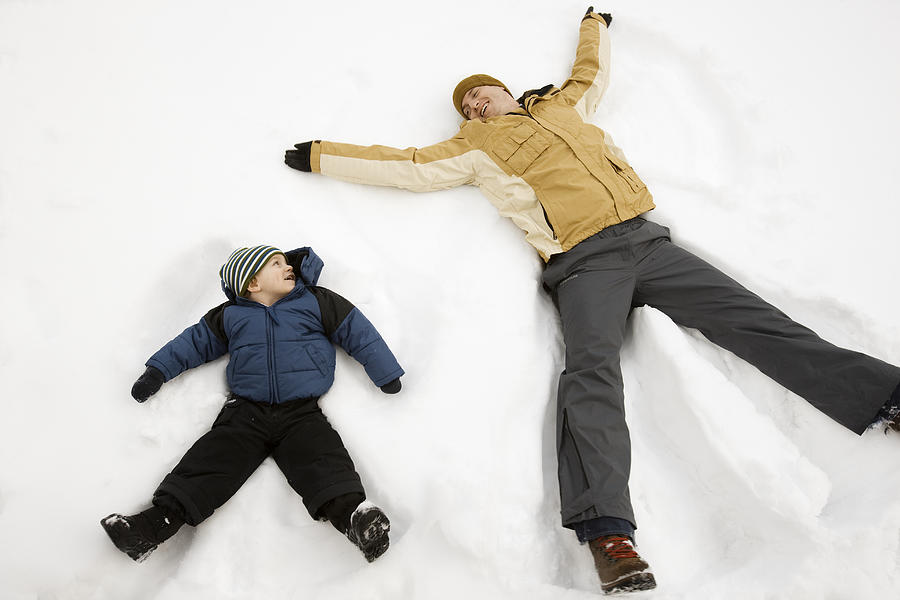 Two people, a man and a child lying in the snow make snow angel shapes.  Photograph by Mint Images