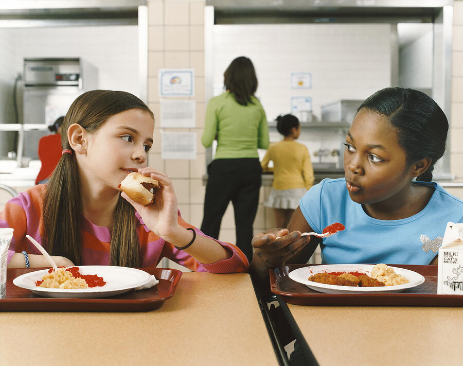 Two Schoolgirls Sitting at a Table in a Canteen Eating Their Lunch Photograph by Digital Vision.