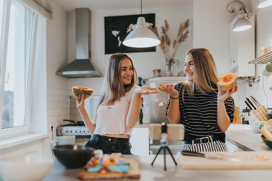 Two teenage girlfriends filming a music video in the kitchen with smart phone Photograph by Visualspace