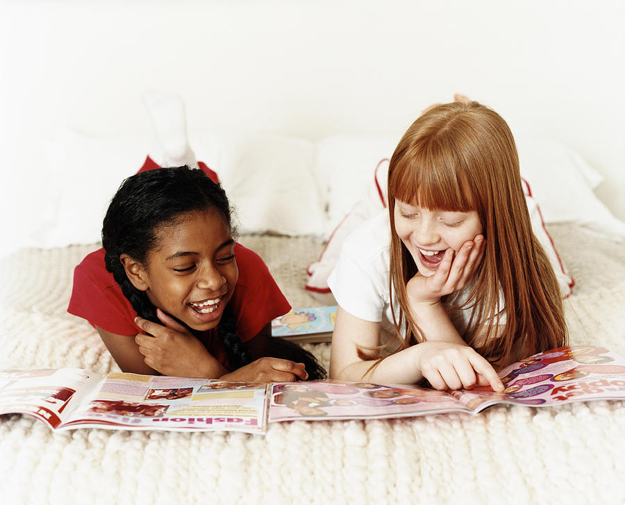 Two Teenage Girls Lying on Bed Reading Magazines, One Girl Pointing at an Image Photograph by Lottie Davies