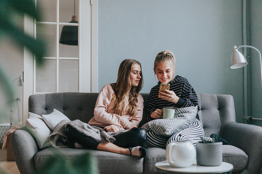 Two teenage girls using smart phone at home on the couch Photograph by Visualspace