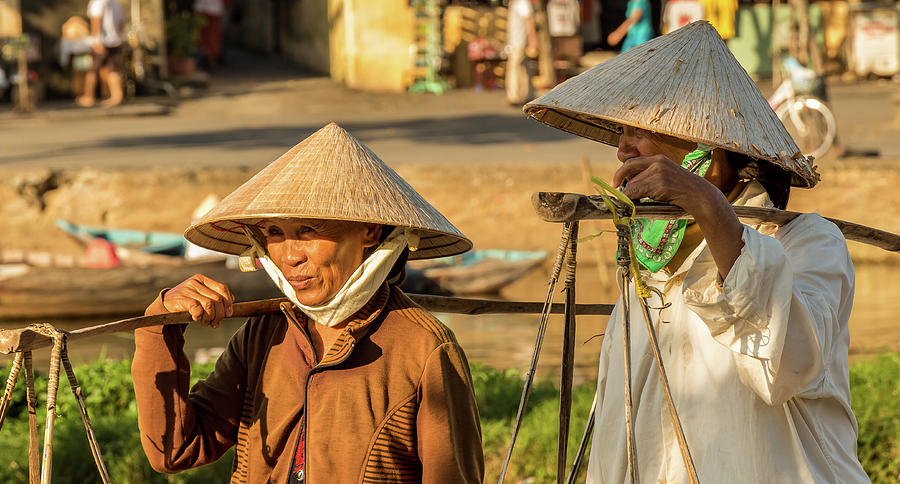 City Photograph - Two Vietnamese women with conical hats, carrying shoulder poles by a river in afternoon, Hoi An, Vietnam - January 10th, 2015 by Snap-T Photography