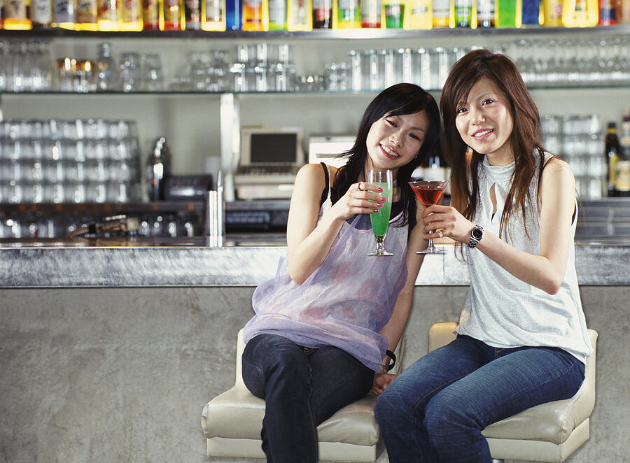 Two Women Sitting at a Bar Making a Toast With Cocktails Photograph by Digital Vision.