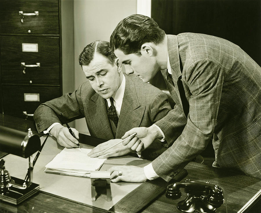 Two young businessmen talking at small desk, (B&W) Photograph by George Marks