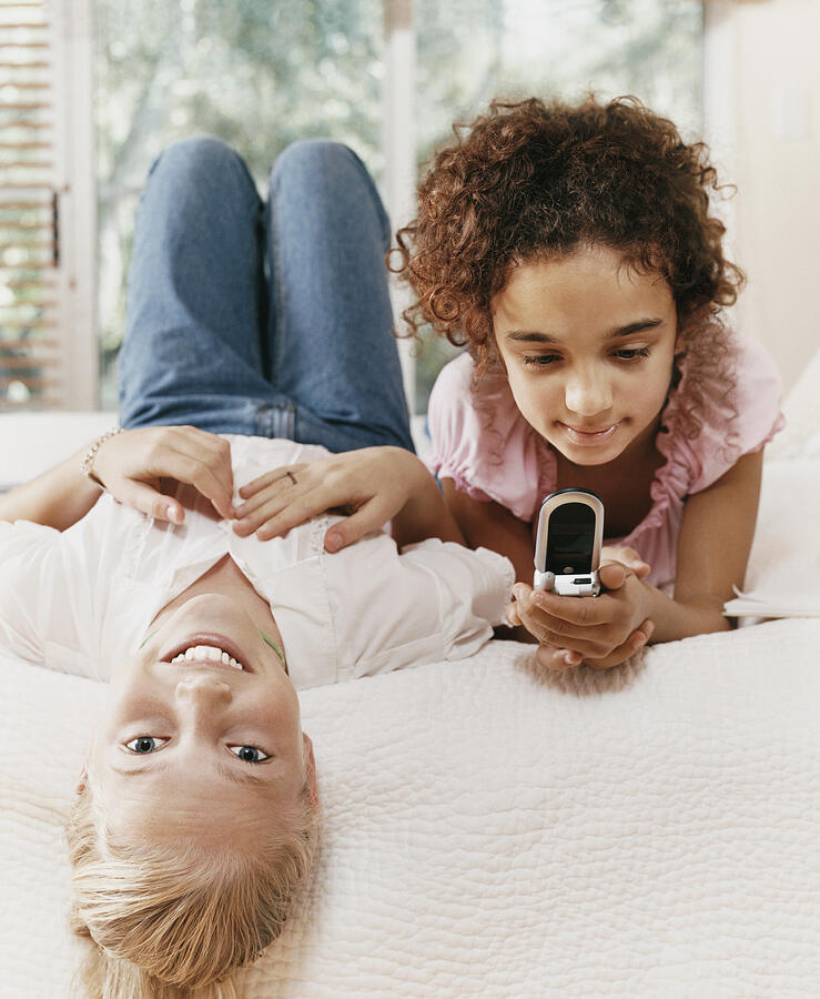 Two Young Girls Lie Side by Side on a Bed, One Texting on Her Mobile Phone Photograph by Digital Vision.
