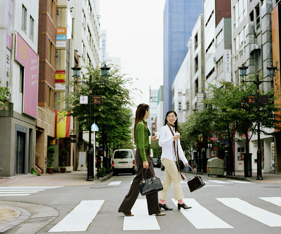 Two young women crossing urban street, having conversation, side view Photograph by Ryan McVay