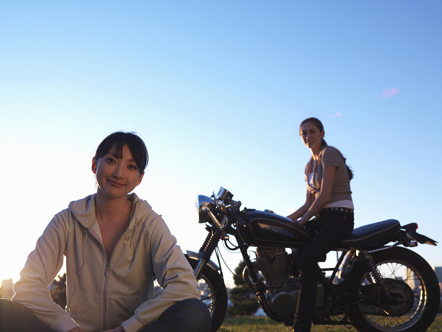 Two young women, one sitting on lawn, one sitting on motorcycle Photograph by Michael H