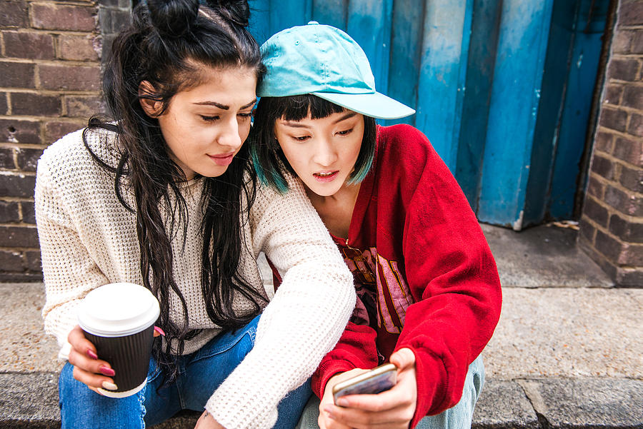 Two young women sitting on sidewalk looking at smartphone Photograph by Bonfanti Diego