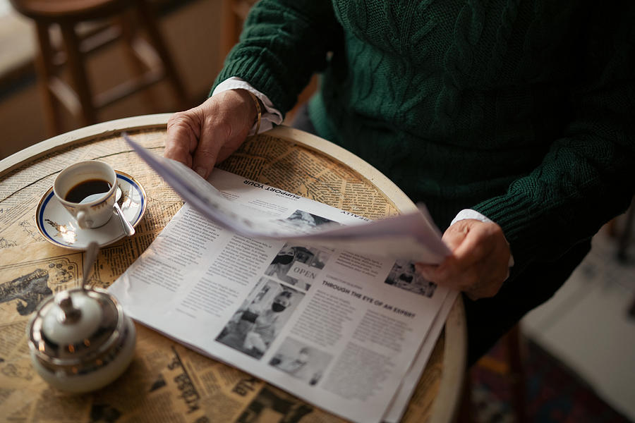 Unrecognizable senior woman reading newspaper and having coffee indoors in café. Photograph by Halfpoint Images
