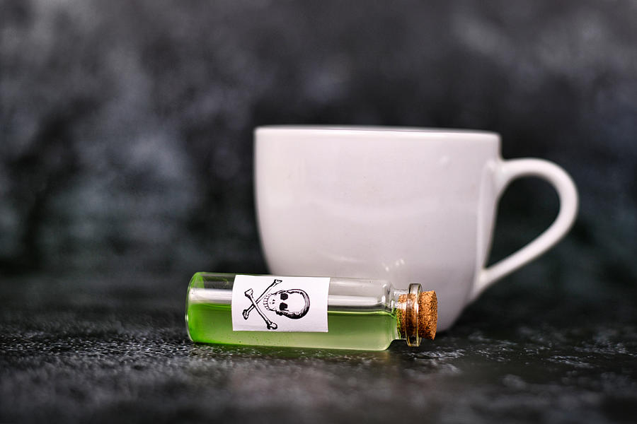 Vial with green liquid and poison skull label in front of blurry tea cup on dark background Photograph by Firn