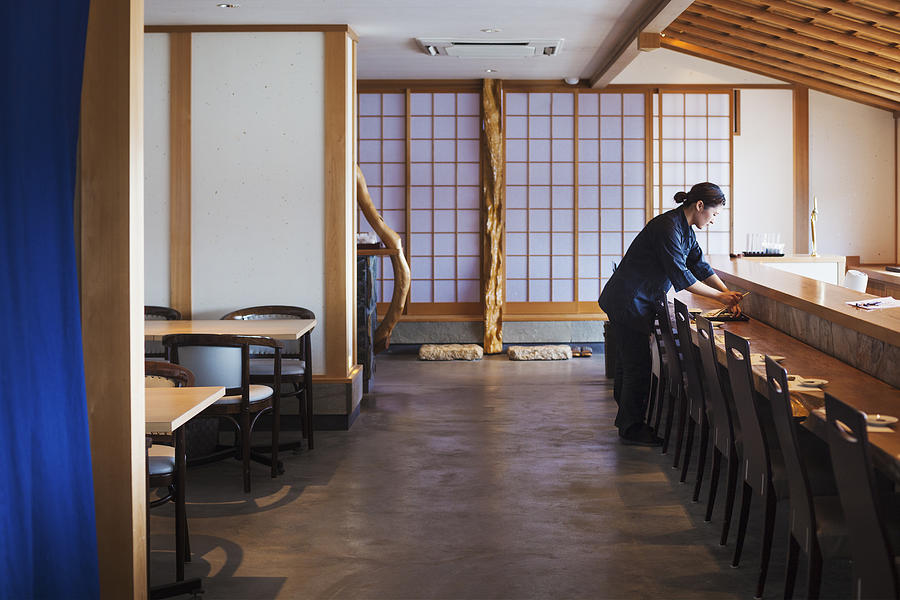 Waitress standing at a counter in a Japanese sushi restaurant, preparing place settings. Photograph by Mint Images