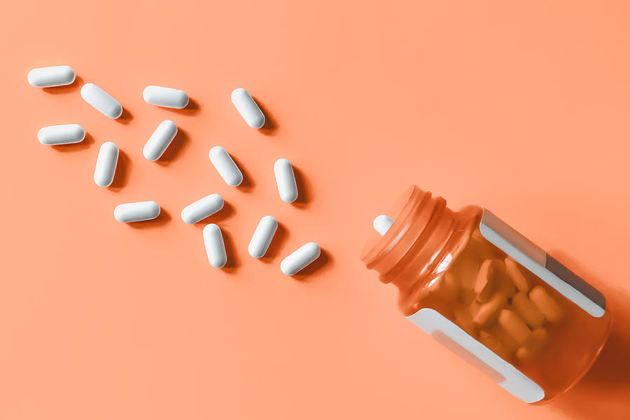 White pills spilling out of prescription bottle onto orange surface Photograph by Grace Cary
