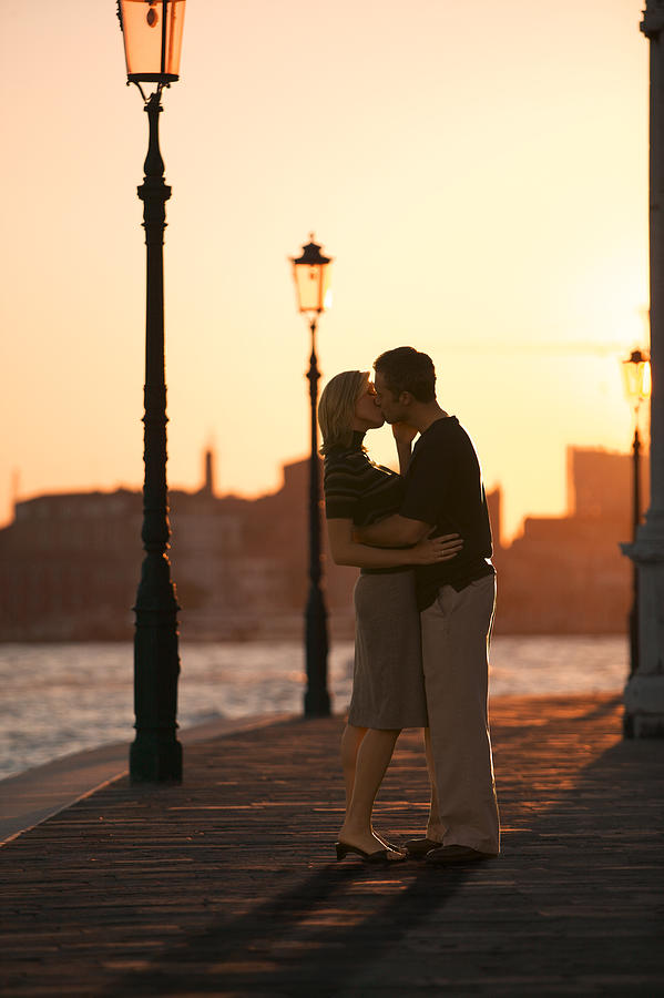 Wide Shot Of A Young Adult Couple As They Embrace And Kiss On A Pier At Sunset Photograph by Photodisc