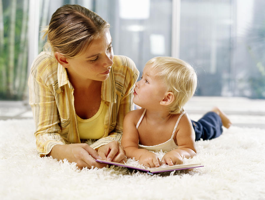 Woman and female toddler (21-24 months) lying on rug with book, looking at each other Photograph by Kraig Scarbinsky