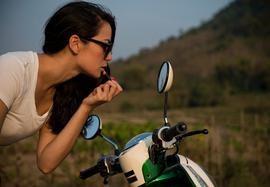 Woman applying lipstick in wing mirror of moped Photograph by Ben Pipe Photography