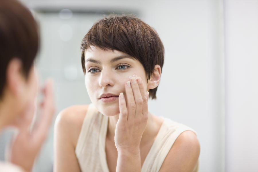 Woman applying moisturizer in mirror Photograph by Zero Creatives