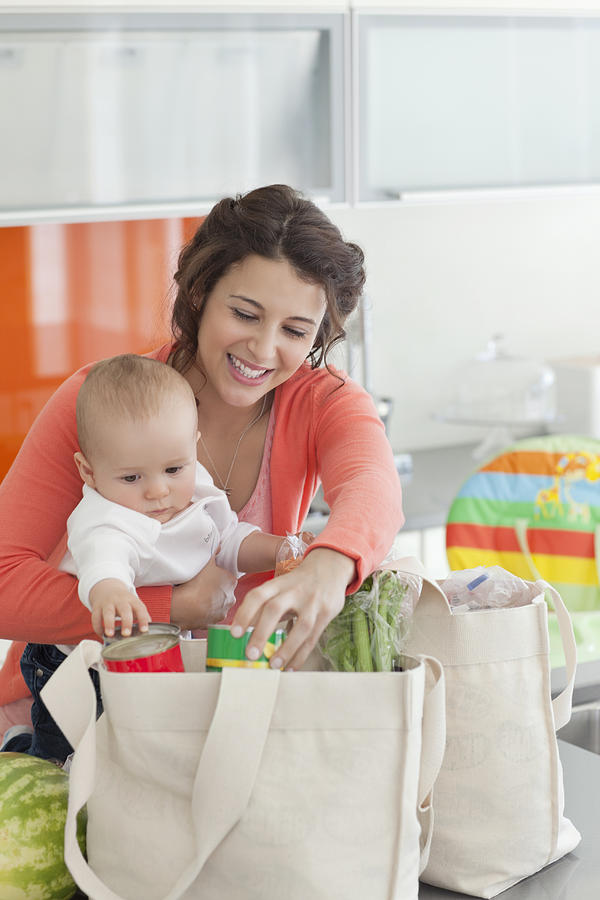 Woman holding baby and unloading groceries from reusable bag Photograph by Sam Edwards