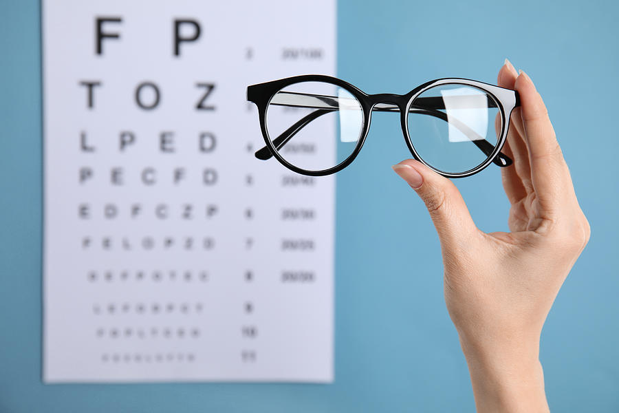 Woman holding glasses against eye chart on blue background, closeup. Ophthalmologist prescription Photograph by Liudmila Chernetska