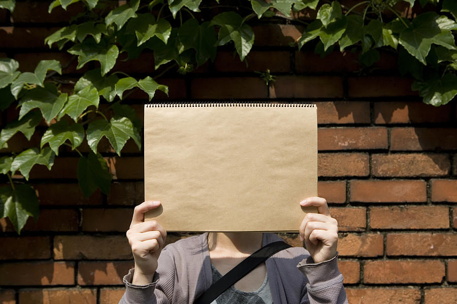 Woman holding sketch pad over face, standing in front of brick wall Photograph by Ichiro