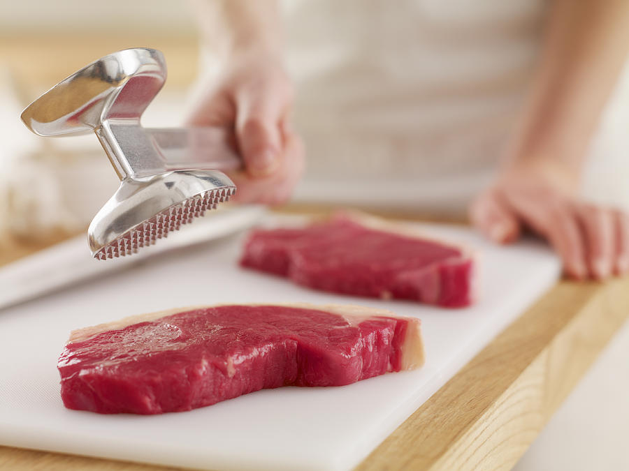 Woman holding tenderizer over raw steaks Photograph by Adam Gault
