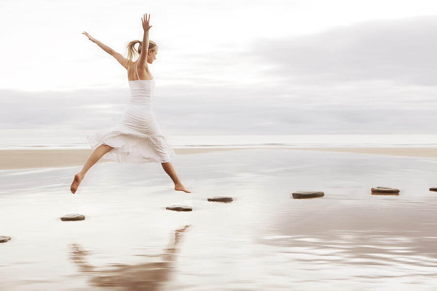 Woman in white dress jumping over stepping stones Photograph by Peter Cade
