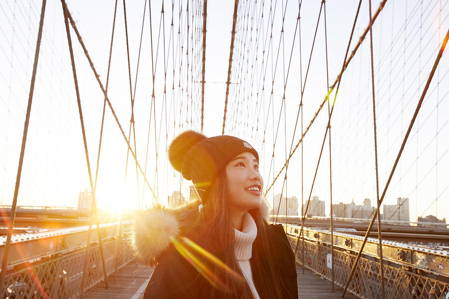 Woman in winter clothing smiling and looking up with sun in background on Brooklyn Bridge Photograph by Chris Tobin